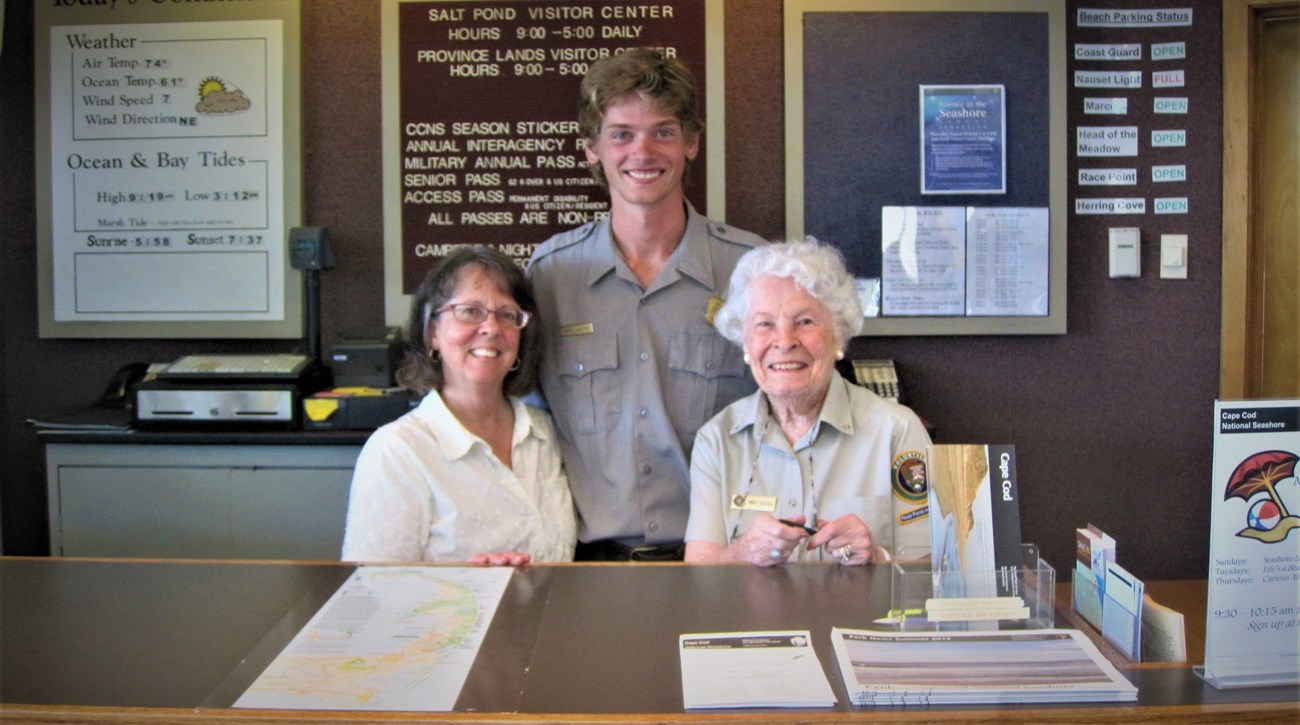 Two volunteers pose with a ranger in between as they stand behind an information desk.
