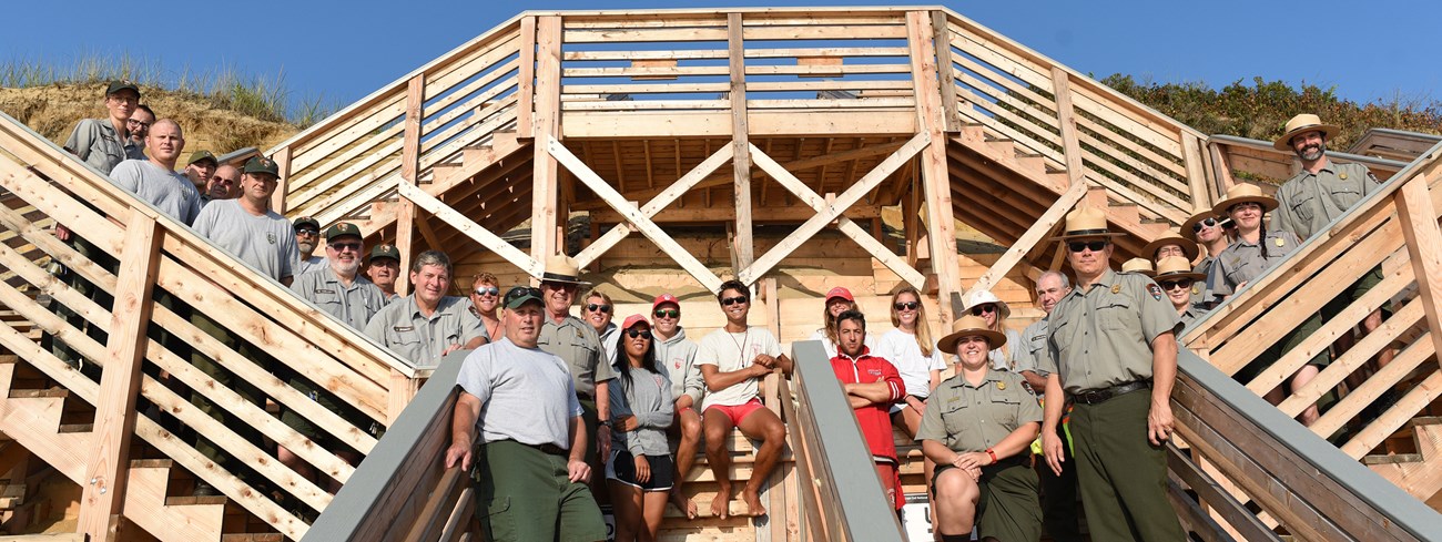 A large group of rangers, and a variety of other National Park Service employees stand on a wooden double staircase leading down a sandy bluff to the beach.