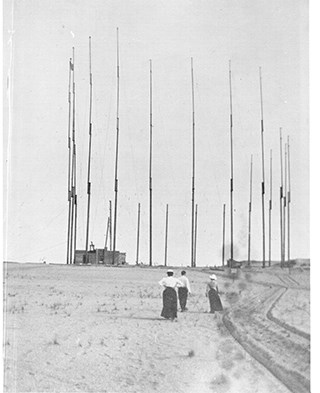 Original Towers A black and white photo of a man and two women standing in an open area facing a building next to a tall circular array of thin antennas.