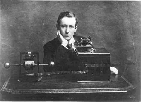 A black and white photo of a young man in a dark suite with a white shirt and dark tie. He rests his head on his hand with his arm supported by a table.