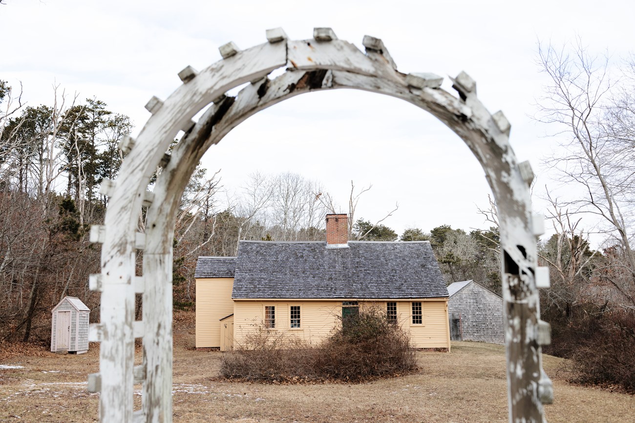 A small yellow one-story house is viewed through a wooden gated archway.