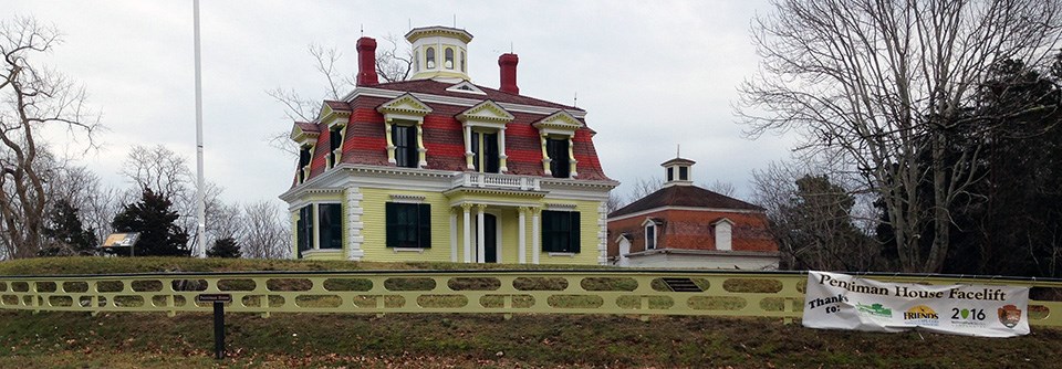 A bright yellow house with white trim sits a top a grassy hill surrounded by trees. There is an ornate wooden fence in the same colors surrounding the house.