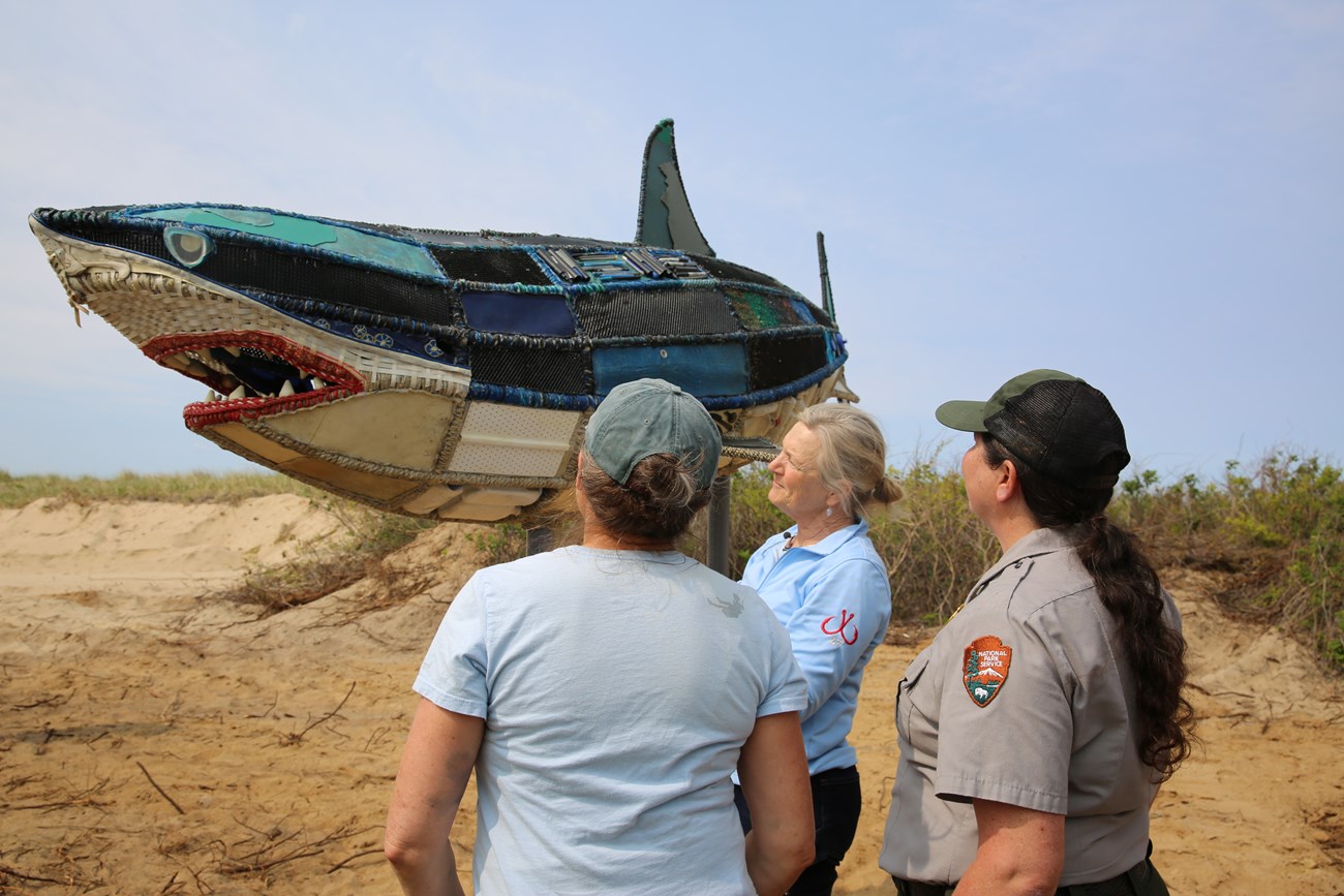 Three people stand in front of a 14 foot shark sculpture made out of marine debris.