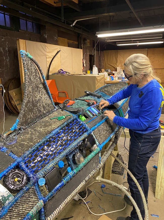 A person secures marine debris to a metal frame that is shaped like a shark.
