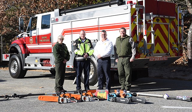 Four people stand in front of a large off-road firetruck. Fire fighting equipment is spread out in front of them.