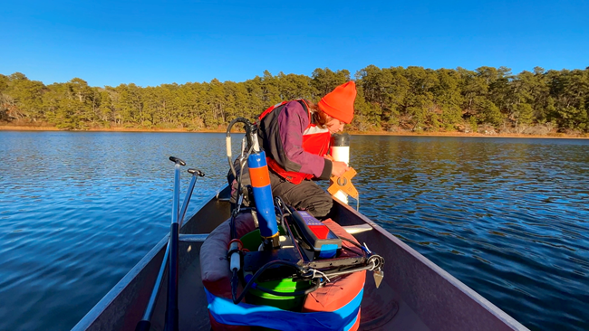 A scientist sits on a canoe on a pond holding an instrument in the water.