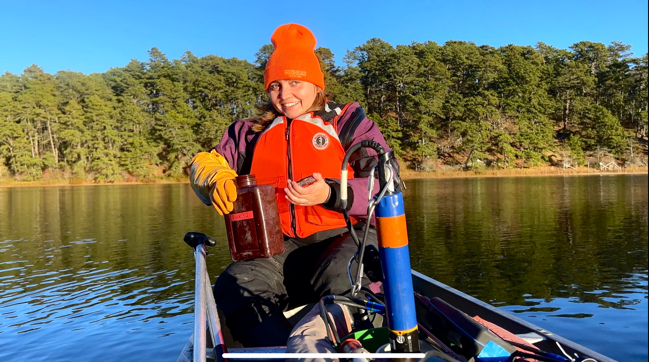 Female scientists sits on a canoe on a large pond holding a plastic container.