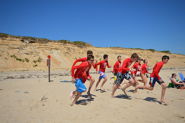 Kids in a variety of  colorful beach gear run down a beach on a sunny day with a cloudless sky.