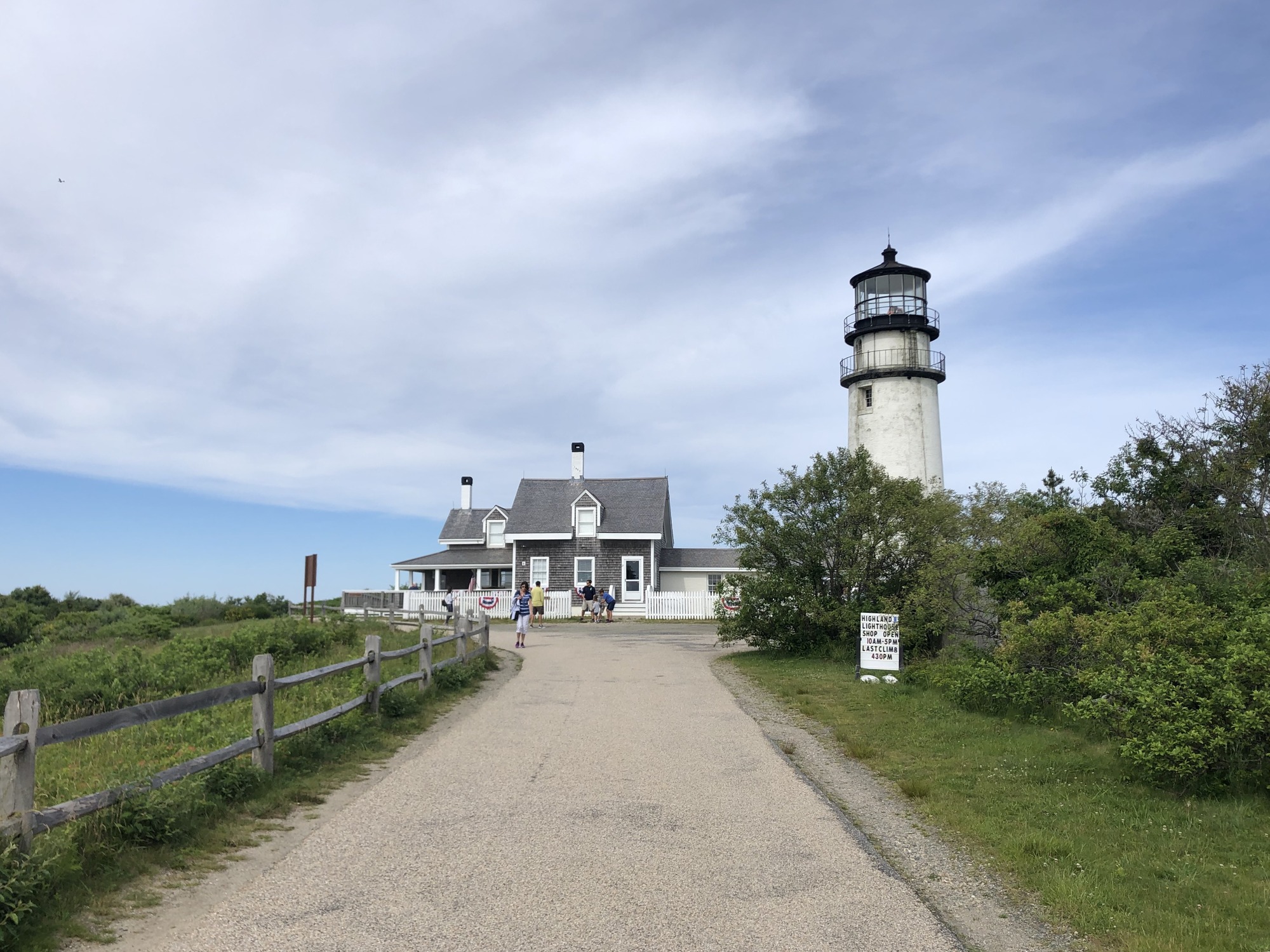 A tall white masonry lighthouse stands tall above a wood-shingled building. The tower shows signs of deteriorated bricks.