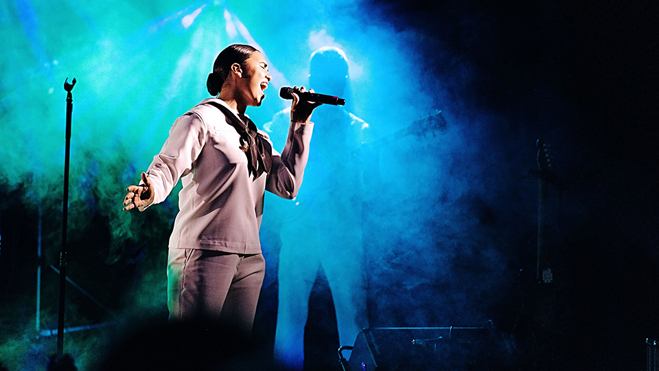 A woman wearing a white United States Navy uniform sings into a microphone in a dark foggy room.