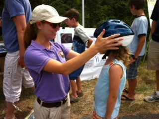 A woman in a blue shirt and a ball cap adjusts a bicycle helmet on a young girl's head.