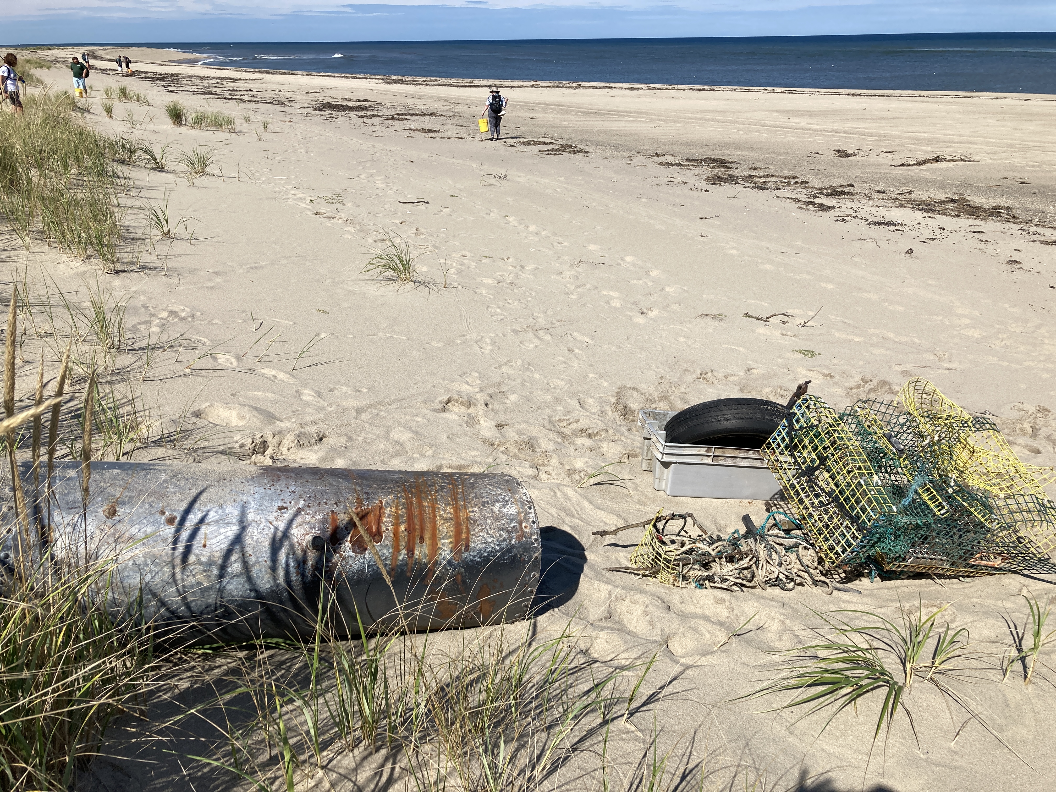 Debris including tires, boxes, and netting sit on a beach. The ocean is in the background. A visitor walks on the beach cleaning up debris.