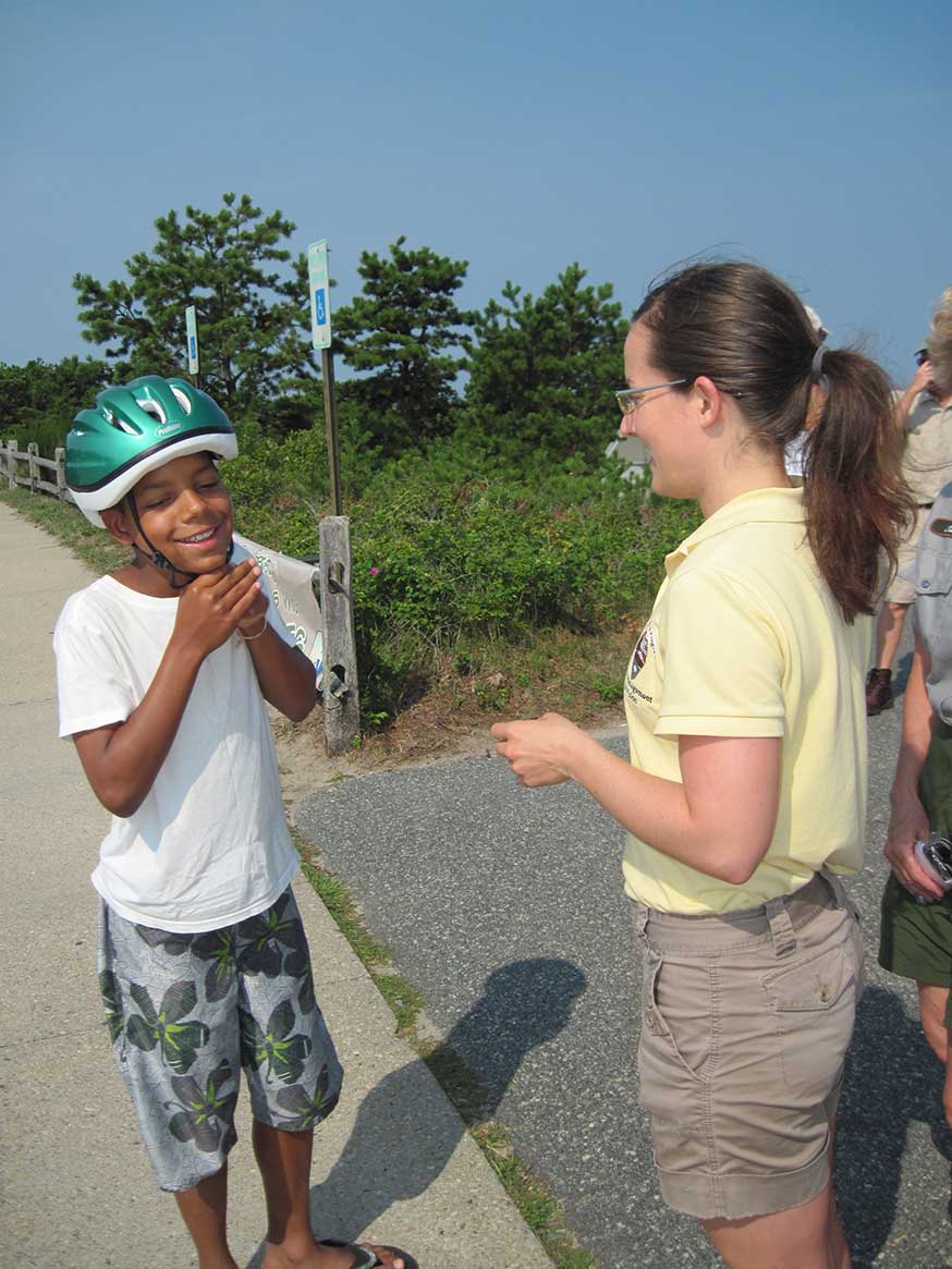 A National Park Service volunteer assists a young visitor in beach clothing with adjusting a bike helmet.