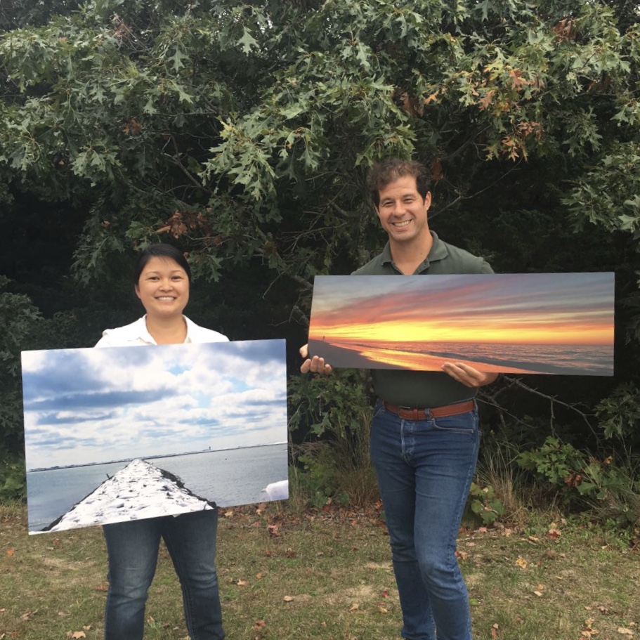 CCNS Photographs by Kekoa Rosehill and Katie Trudeau on Exhibit at Salt Pond Visitor Center. A man and a woman stand in the grass in front of a large bush holding photographs printed on large metal sheets.