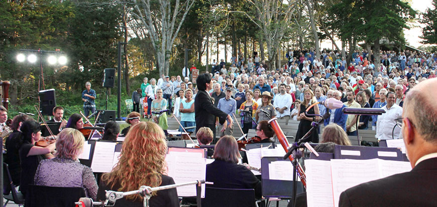 Looking out from behind a symphony playing out doors to a large crowd sitting on wooden benches.