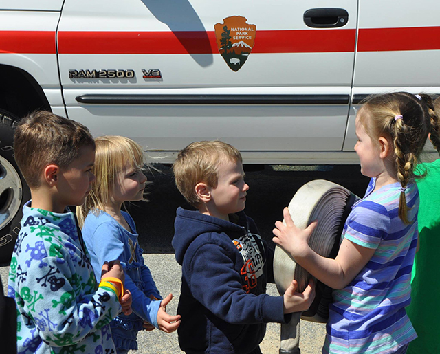 A young girl with her hair in two high braided pigtails wearing a striped blue shirt hands a rolled fabric fire hose to a smaller fair haired boy in a dark blue sweatshirt. Two more children stand in line behind the boy.