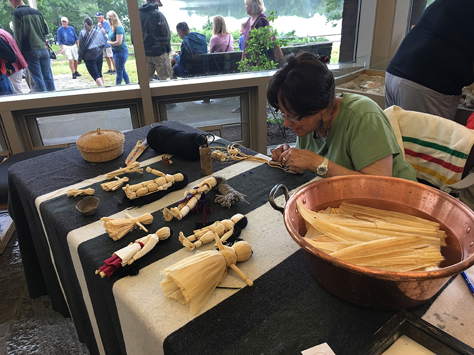 A woman makes dolls out of corn husks at a table in front of a large glass window.