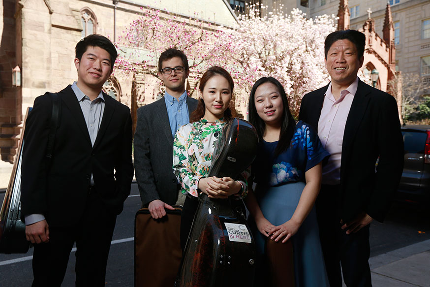 Five musicians pose in formal wear with instruments.