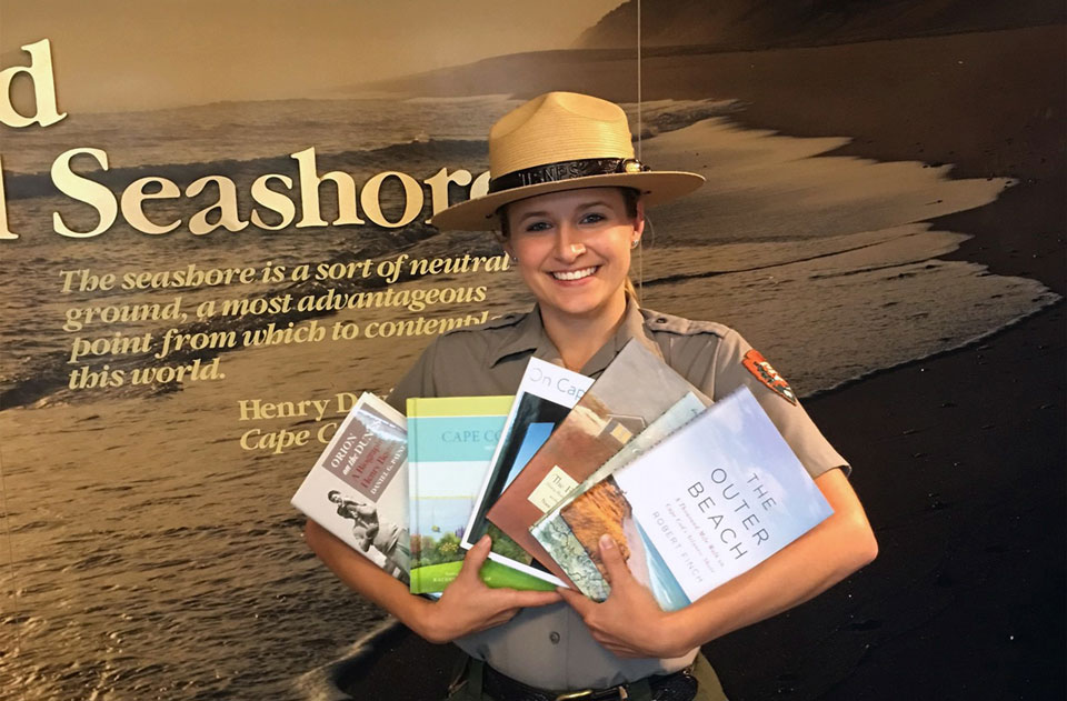 A ranger wearing a gray shirt and wearing a flat straw hat holds books fanned out in her arms.