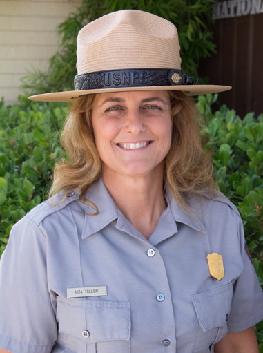 A woman in a grey ranger shirt and a straw ranger hat smiles.