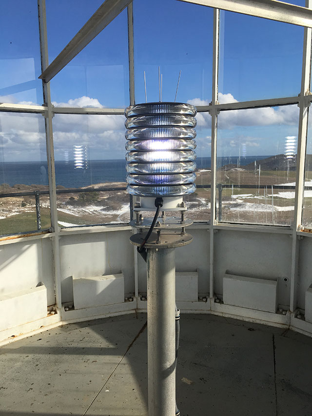 A blue LED light sits on a post inside the glass room of a lighthouse tower. Through the glass, a blue sky with light fluffy clouds can be seen.