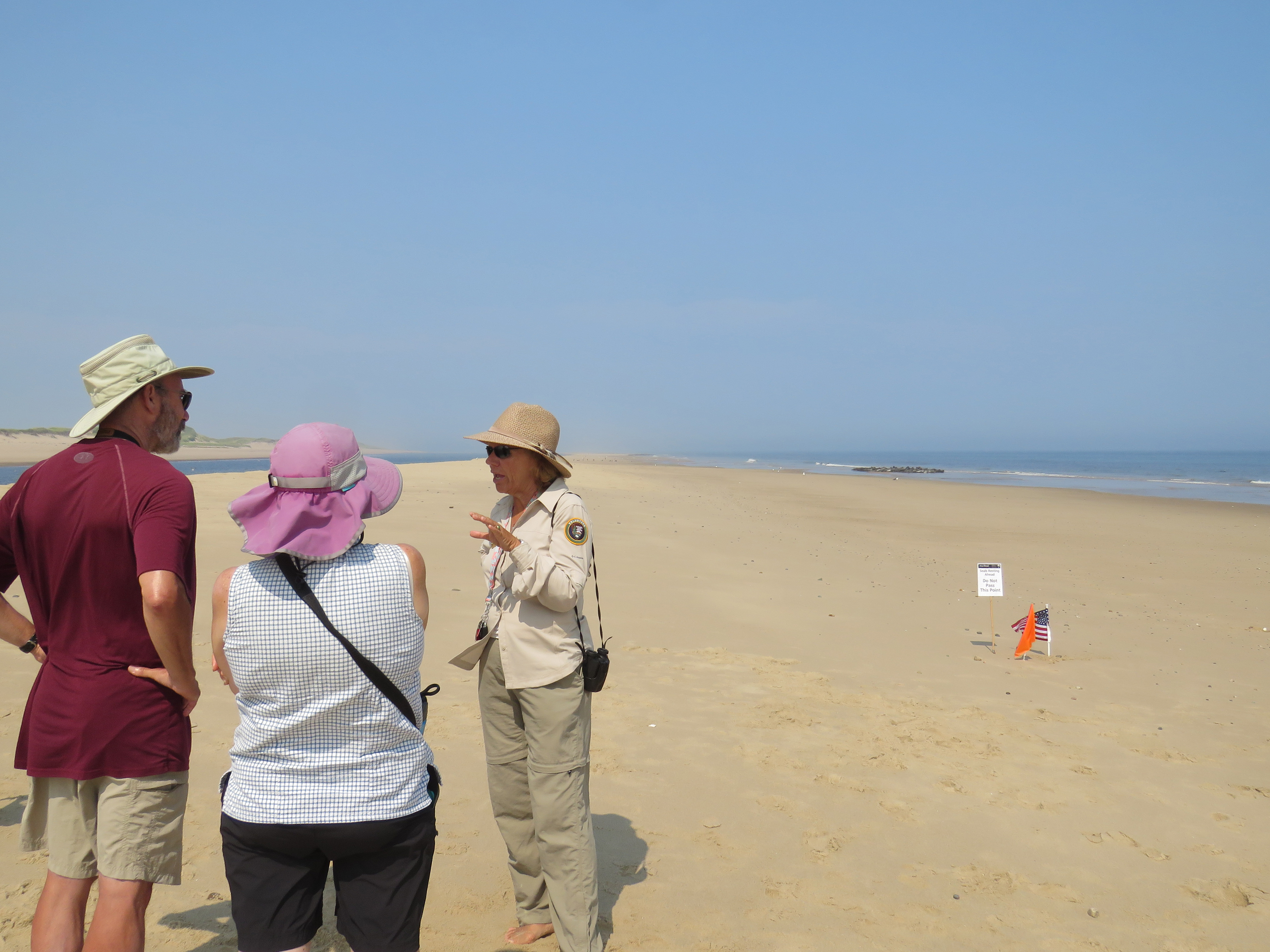 A woman talks to two visitors on a sunny beach. Behind her is a sign telling people to keep back, flanked by orange flags.