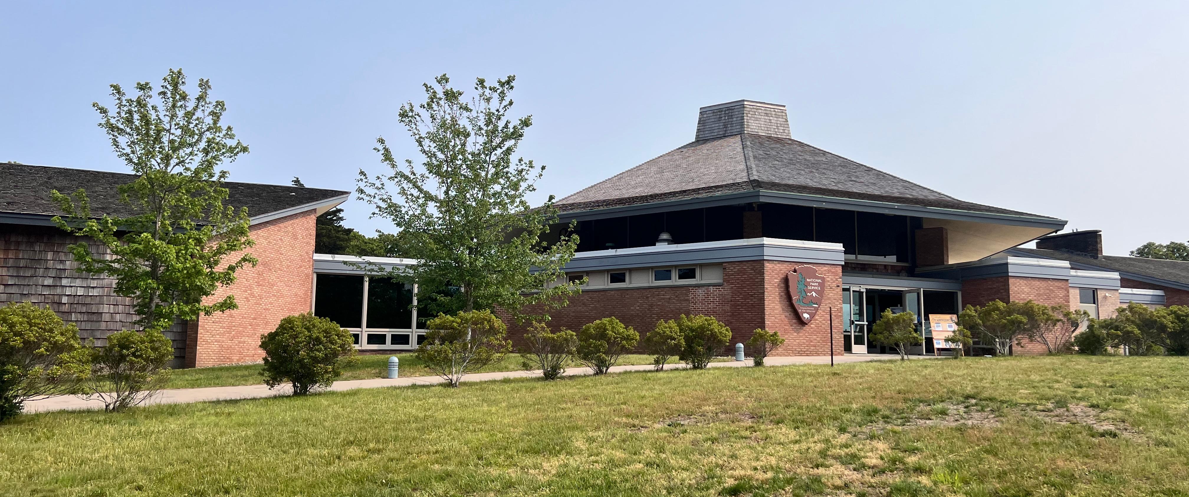 A brick and tan shingled visitor center with a green lawn and bushes.