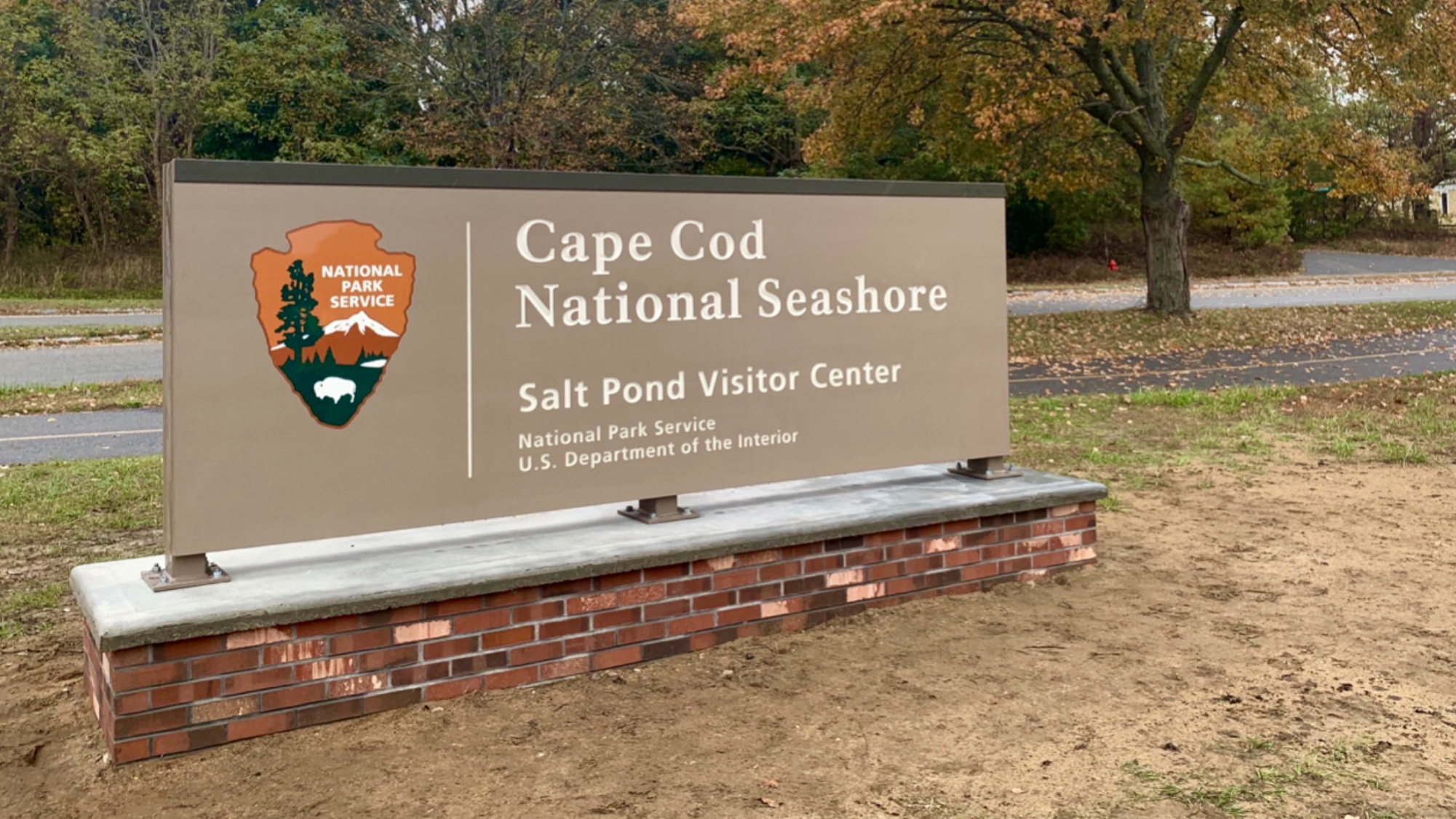 A new, brown park entrance sign sits on top of several rows of bricks. The sign reads: Cape Cod National Seashore, Salt Pond Visitor Center. There is a National Park Service arrowhead on the sign.