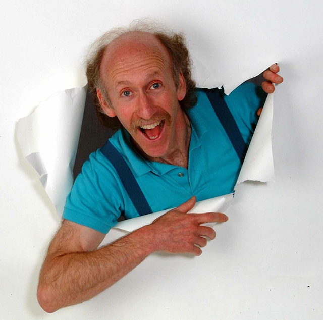 A man with slightly balding hair wearing a blue polo shirt and suspenders breaks through a white paper wall.