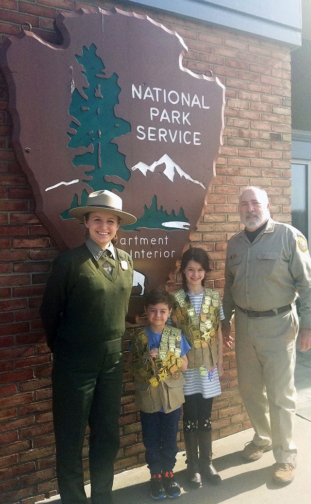 A ranger, and a volunteer in a tan shirt stand on either side of two children wearing vests covered in plastic badges.