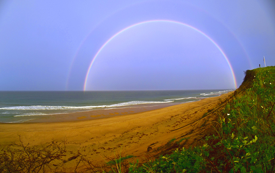 Karst Hoogeboom, Multiple Pots of Gold Looking out over a sandy beach from atop a bluff. A wide double-rainbow arches through the sky.