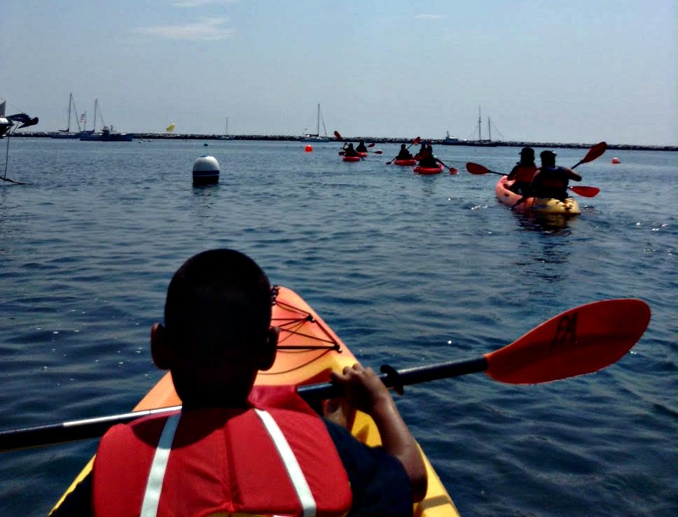 A young man at the end of a line of kayaks, paddles across still water.