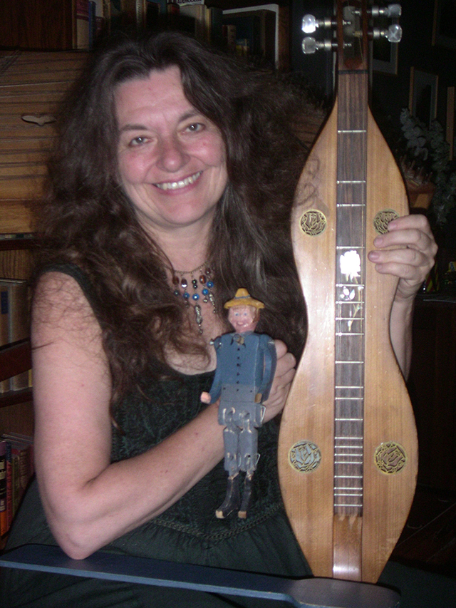 A woman with long dark hair sits with a wooden stringed instrument in her lap.