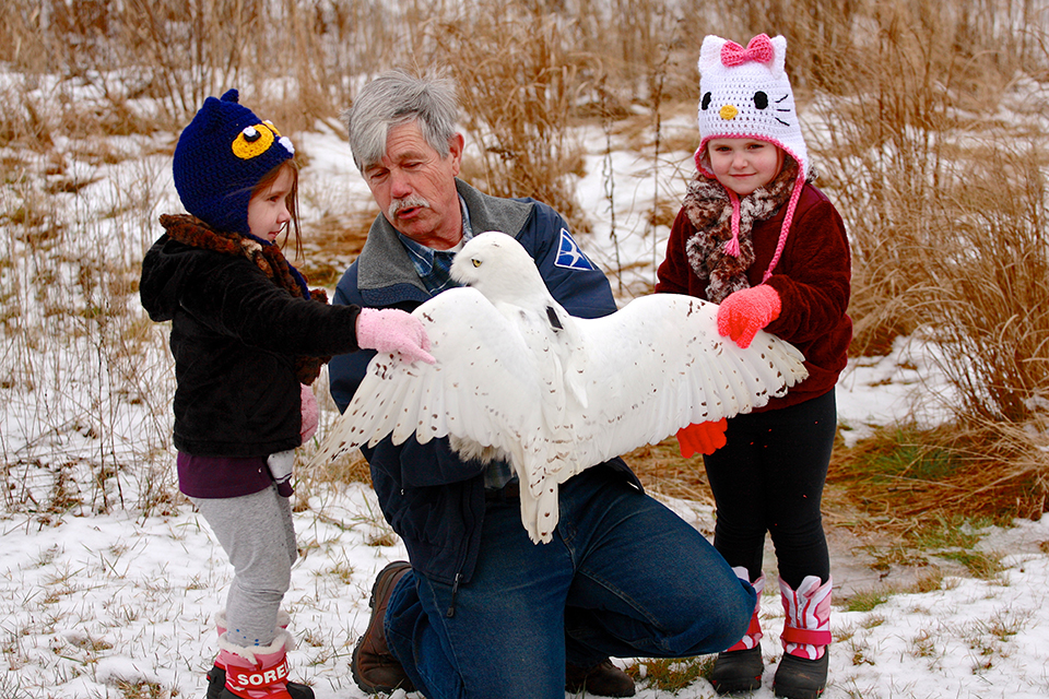 A man with gray hair and a mustache holds a large white owl with its wings spread as two children watch.