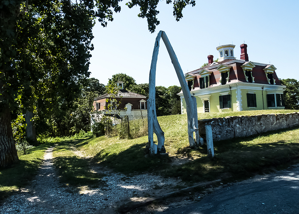 A bleached white whalebone arch rises above a pathway leading up to a bright yellow house.