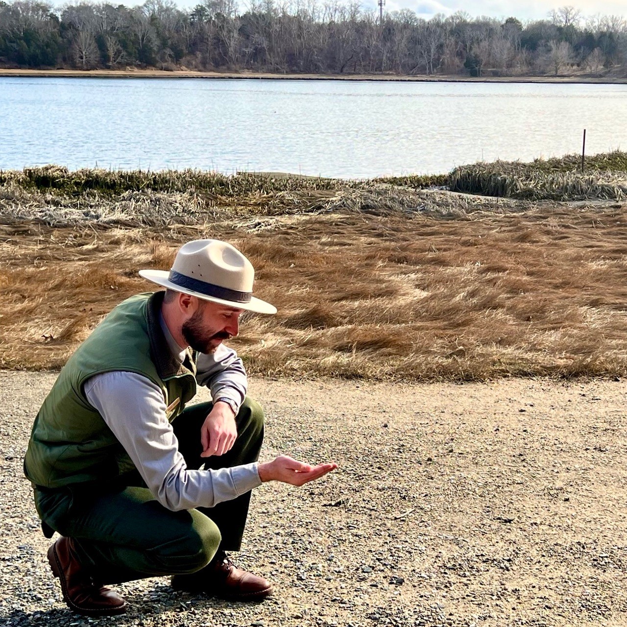 Ranger at Salt Pond, NPS A man kneels on the ground and cups his hand to show off an object. A pond lies in the background.