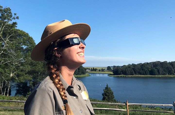 A ranger with long braided hair and a flat wide-brimmed straw hat wears cardboard sunglasses and looks up at the sky.