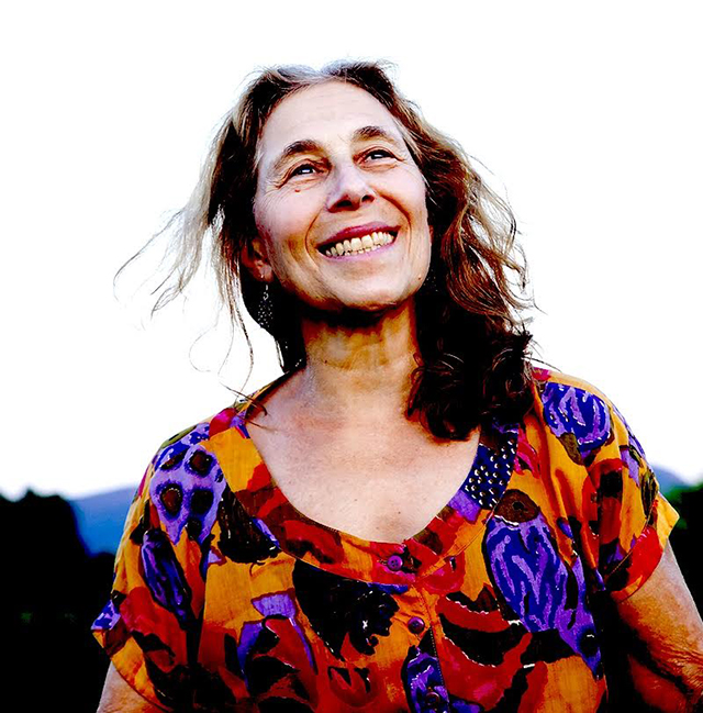 A woman with long dark hair, and a bright multi-colored top looks up and smiles.