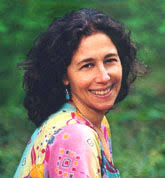A woman with dark curly hair, wearing a brightly colored shirt, sits outdoors on a sunny day.
