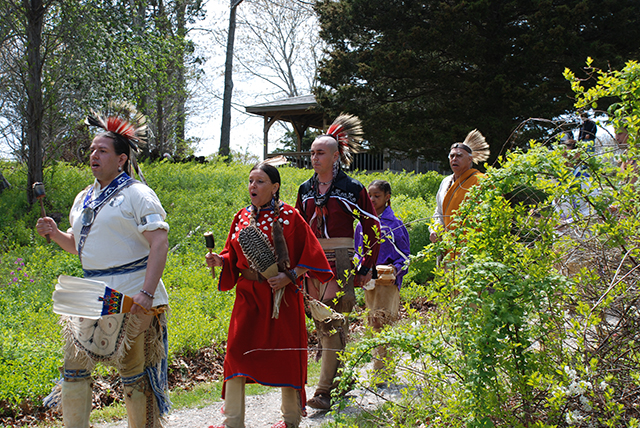 Native Americans in full dress walk down a concrete pathway from behind bushes, singing and chanting.