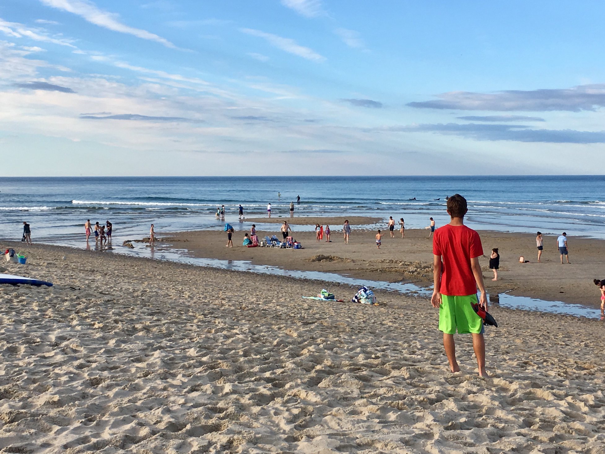 Summer evening at Coast Guard Beach. Man in red shirt looks out to the ocean waves lapping up on the beach. A blue sky is dotted with thin, wispy clouds. People sit and stand along the beach.