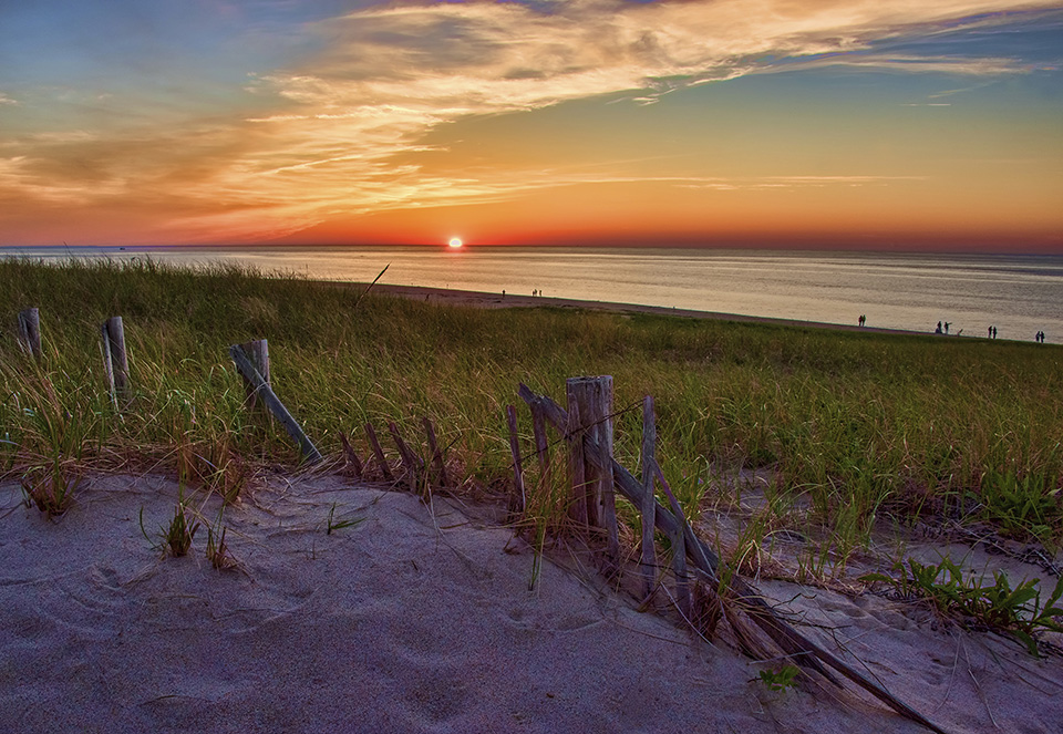 A photograph of a setting sun dropping behind a glowing ocean and reflecting off of the clouds.