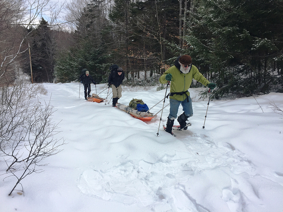 Three people wearing heavy winter clothing, snow shoes, and using trekking poles pull two sleds of equipment through the snow.
