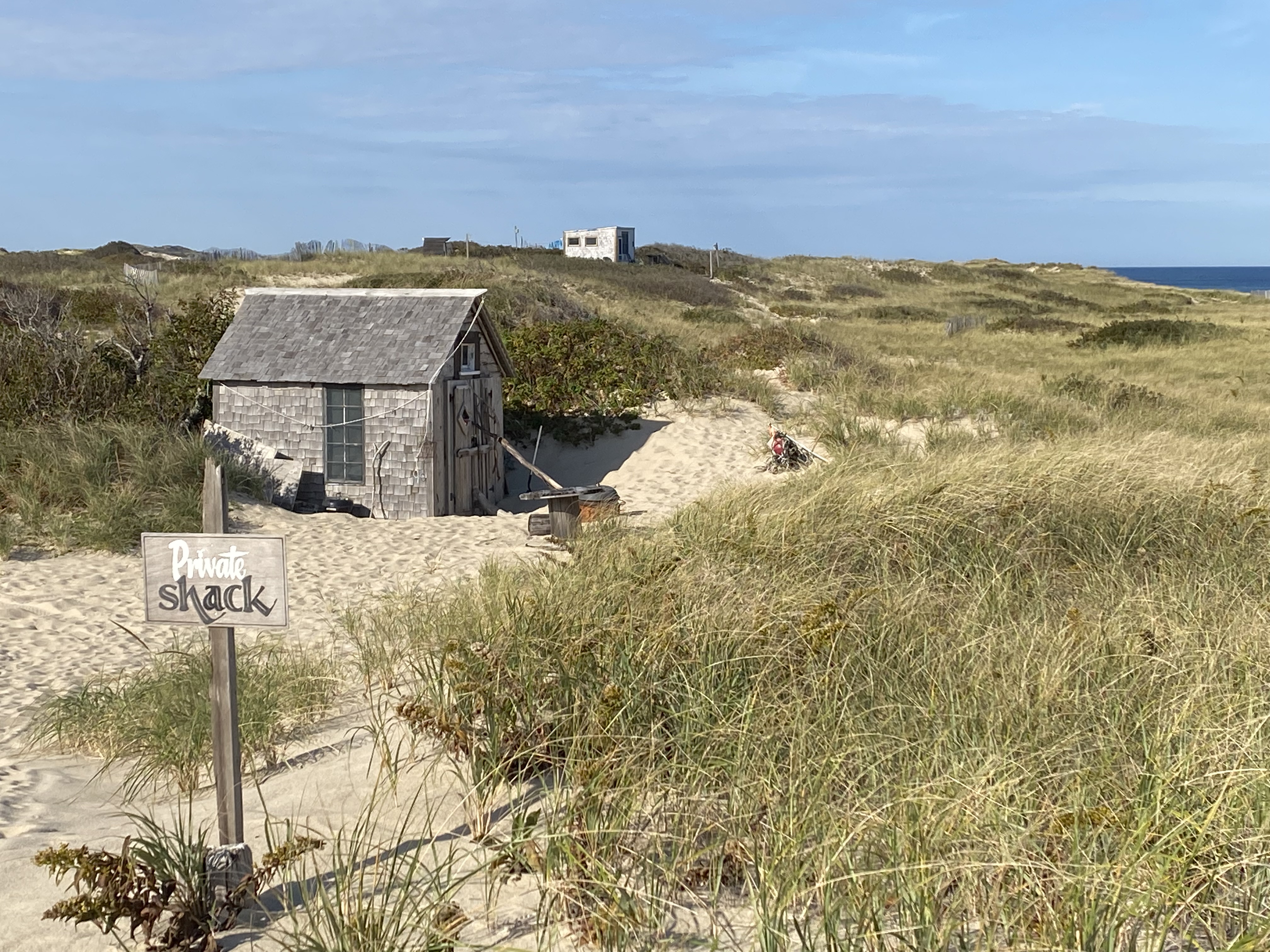 A dune shack sits among rolling sand dunes against a blue sky.