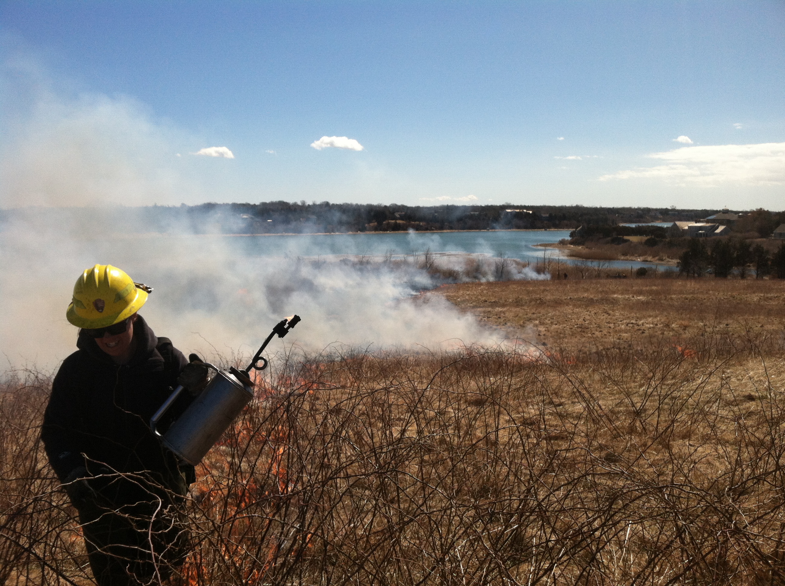 prescribed burn fort hill person in yellow hard hat holding a mental gas can walks along a meadow