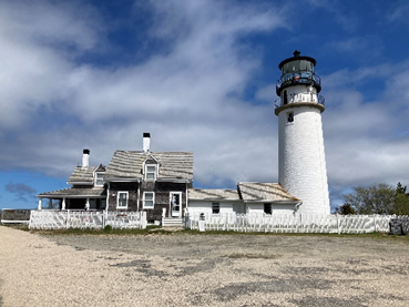 Tall white light house stands next to a cottage with white picket fence against a blue sky.