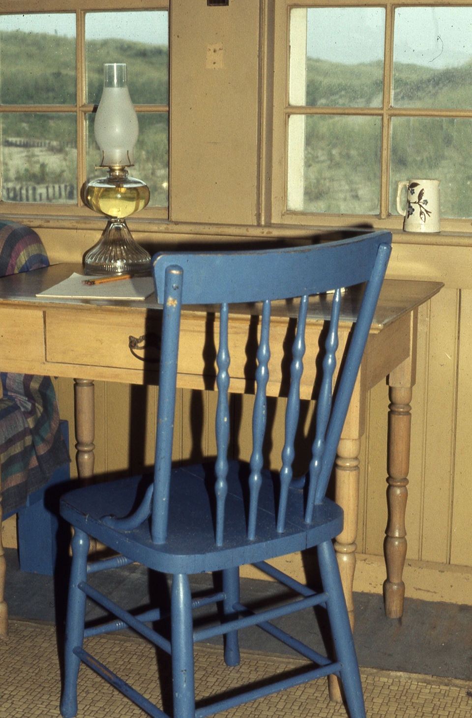 A chair sits at a small wooden table with a lamp on it. Behind the table is a window looking out to grassy dunes.