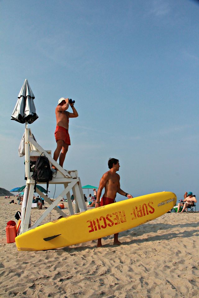 Two lifeguards, one on a tall stand looking at the water with binoculars, the other carrying a yellow surfboard