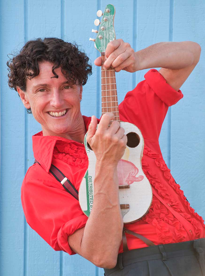 Female musician with brown curly hair and a bright red shirt smiles and holds up an ukulele.
