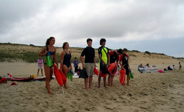 Junior lifeguards stand ready on an overcast day on a beach to practice their skills.
